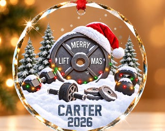 Könnte beinhalten: Ein runder Weihnachtsbaumschmuck aus Glas mit einer Hantelscheibe, die eine Weihnachtsmütze trägt, umgeben von Schnee, Tannen, Kurzhanteln und Kettlebells. Der Text lautet MERRY LIFT MAS, CARTER und 2026. Er hängt an einem roten Band.