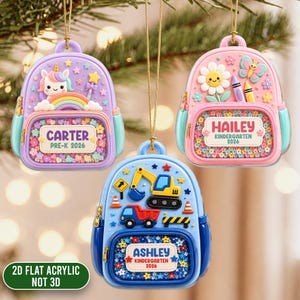 May include: Three backpack-shaped ornaments hanging by gold strings. The purple one features a unicorn, rainbow, and the text CARTER PRE-K 2026. The pink one has a flower, butterfly, and HAILEY KINDERGARTEN 2026. The blue one shows construction vehicles and ASHLEY KINDERGARTEN 2026.