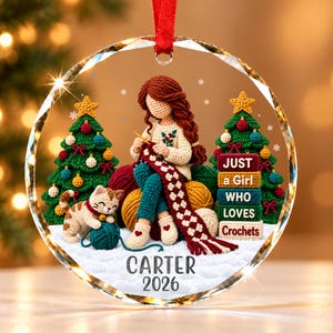 May include: A round glass Christmas ornament featuring a crocheted figure with long brown hair, a cream sweater, and blue pants, sitting between two green crocheted Christmas trees. A small cat plays with yarn. Text reads: JUST a Girl WHO LOVES Crochets, CARTER, 2026.