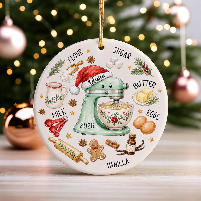 May include: A round white ceramic ornament featuring a light green stand mixer wearing a red Santa hat with the name Olivia and the year 2026. Illustrations of baking ingredients like flour, sugar, butter, eggs, milk, and vanilla surround the mixer.