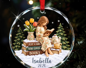 Könnte beinhalten: Ein kreisförmiger Glasschmuck mit rotem Band, der ein Mädchen zeigt, das auf einem Stapel Bücher mit der Aufschrift Just a Girl Who Loves Books liest. Die Szene umfasst gelbe und orangefarbene Tulpen, eine Teekanne, eine Teetasse und Tannenbäume auf Schnee, mit dem Namen Isabella und dem Jahr 2026 am unteren Rand.
