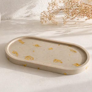 Peut inclure: Plateau ovale en béton beige clair avec des accents de feuilles d'or. Le plateau a une surface lisse et des bords arrondis. Un objet décoratif pour ranger de petits objets.