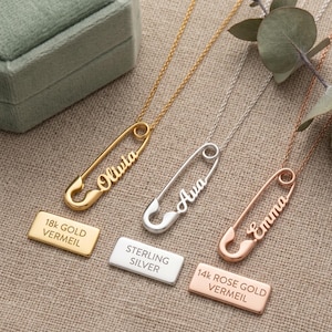 Könnte beinhalten: Drei personalisierte Sicherheitsnadel-Halsketten in Gold, Silber und Roségold. Jede Halskette trägt einen Namen: Olivia, Ava und Emma. Das Bild zeigt auch drei Metallanhänger mit den Materialien: 18 Karat Gold Vermeil, Sterlingsilber und 14 Karat Roségold Vermeil.