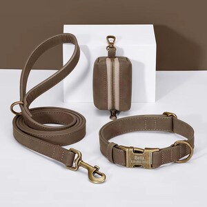 Pode incluir: Um conjunto de acessórios para cães em verde-oliva suave. O conjunto inclui uma trela, coleira e dispensador de sacos para resíduos. A coleira tem uma fivela de metal com o nome "Bella" e um número de telefone. Todos os itens têm ferragens de metal a condizer.