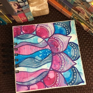 May include: A watercolor painting of a mandala with pink, blue, and purple colors. The mandala is made up of circles, dots, and lines. The painting is on a white background.