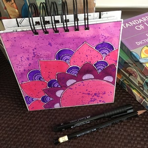May include: A pink and purple watercolor painting of a mandala design in a spiral notebook. The notebook is open and the painting is on the right page. The notebook is on a brown surface with two black pens.