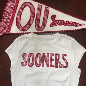 May include: White t-shirt with "SOONERS" in red gingham letters. A pennant with "OU" in red gingham and "Sooners" in script, with red and white trim. The pennant is white with red gingham letters.