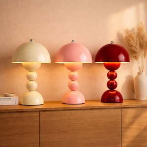 Puede incluir: Tres lámparas de mesa con forma de hongo en crema, rosa y rojo. Cada lámpara tiene una pantalla redondeada y una base compuesta por esferas apiladas. Las lámparas están colocadas sobre una superficie de madera, con un jarrón y una planta decorativa en el fondo.