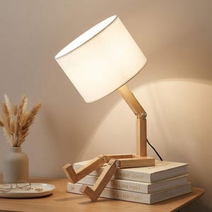 May include: A whimsical wooden desk lamp with a white lampshade. The lamp's design features adjustable wooden legs and a rectangular base, giving it a playful, human-like appearance. The lamp is sitting on top of a stack of books.