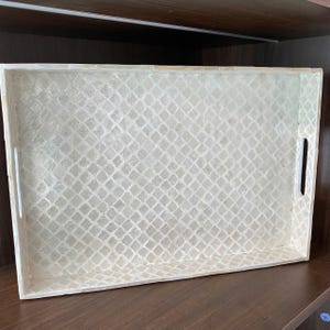 Könnte beinhalten: Ein rechteckiges Serviertablett mit einer cremeweißen, schimmernden Perlmutt-Intarsie in einem sich wiederholenden Vierpassmuster. Das Tablett hat erhöhte Seiten, einen flachen Boden und zwei integrierte vertikale Griffausschnitte an den kurzen Seiten.