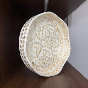 May include: A decorative, off-white serving tray with a leaf-like pattern. The tray's rim is adorned with a repeating pattern of small, light brown shapes. The interior features a detailed design of light brown leaf shapes against a white background.