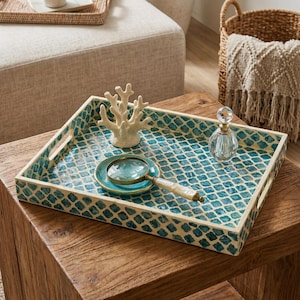 May include: A decorative rectangular tray with a teal and cream mosaic pattern. The tray holds a white coral sculpture, a magnifying glass, and a small glass perfume bottle. The tray has handles and is on a wooden table.