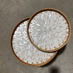 Puede incluir: Dos bandejas decorativas redondas. Cada bandeja presenta un centro blanco con un patrón floral y un borde marrón tejido. Las bandejas están apiladas, creando un efecto en capas. Las bandejas están hechas de materiales naturales.