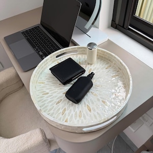 May include: A round decorative tray featuring a white and cream-colored mosaic pattern made of small, petal-shaped pieces. The tray has two integrated handles on opposite sides and holds a black wallet, a black bottle, and a small white canister.