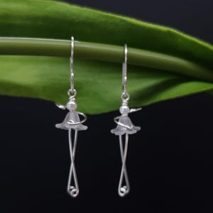 Puede incluir: Un par de pendientes de plata con un diseño floral. Cada pendiente presenta una pequeña forma de flor translúcida con un tallo de alambre plateado que se curva en la parte inferior. Los pendientes se muestran sobre un fondo oscuro con una hoja verde.