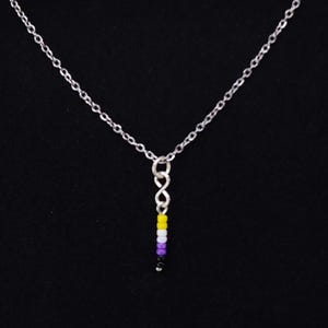 Puede incluir: Un collar de cadena plateada con un colgante de pequeñas cuentas de colores. Las cuentas están dispuestas verticalmente, mostrando amarillo, blanco, morado y negro. El collar se presenta sobre un fondo negro.