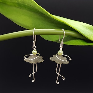 May include: A pair of handmade earrings featuring a ballerina design. Each earring has a gray flower-shaped body, silver wire arms and legs, and a small green bead head. The earrings hang from silver hooks. The background is black with a green stem and leaf.