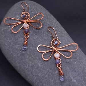 May include: Copper wire dragonfly earrings with blue, purple, and white beads. The dragonfly wings are formed from curved copper wire. The earrings have a hook closure and a teardrop-shaped bead at the bottom.