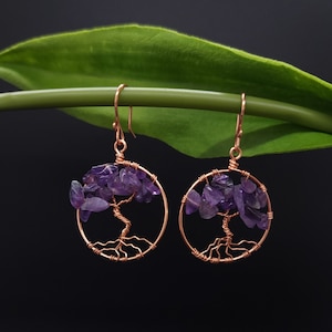 Peut inclure: Une paire de boucles d'oreilles en fil de cuivre. Chaque boucle d'oreille présente un cadre circulaire avec un motif d'arbre et est remplie de petites pierres d'améthyste violettes. Les boucles d'oreilles sont suspendues à des crochets d'oreilles en cuivre.