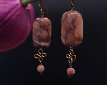 May include: A pair of earrings with rectangular, pink and brown stone beads. The earrings have gold-colored hooks and decorative wirework with a small, round pink bead dangling below. The background is dark, with a purple flower and green stem.