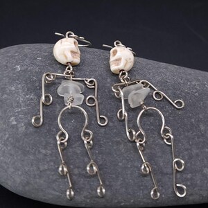 May include: A pair of unique skeleton earrings. Each earring features a small, off-white skull, a clear, irregular-shaped stone, and a silver-colored wire frame forming the body. The earrings are designed with a whimsical, artistic flair.