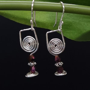 Peut inclure: Boucles d'oreilles en fil d'argent avec un motif de verre à vin. Chaque boucle d'oreille présente une spirale, un groupe de perles violettes et une base. Les boucles sont suspendues à un crochet en argent. Le fond est noir avec une tige et une feuille vertes.