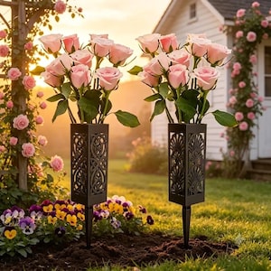 May include: Two black metal garden stakes with square, open-work containers holding bouquets of light pink roses. The left stake features an intricate Celtic knot pattern, while the right stake displays a fan-like, art deco style geometric design.