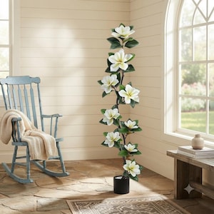 Puede incluir: Un árbol de magnolia artificial alto con un tallo delgado de color marrón oscuro, múltiples flores blancas y hojas de color verde oscuro. La planta está en una maceta cilíndrica negra sencilla, erguida como un acento botánico decorativo.