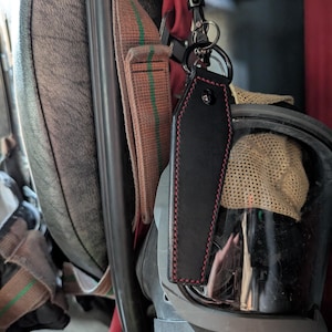 May include: Black leather coffin-shaped keyring with red stitching and a metal ring. The keyring is attached to a black metal clip. The background includes safety equipment and straps.