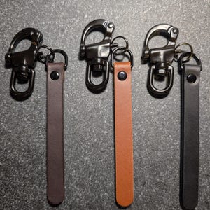 May include: Three leather key fobs with dark metal swivel trigger snaps. Each fob features a flat leather strap in dark brown, tan, or black, secured with a matching dark metal rivet. The hardware includes a circular ring connecting the strap.