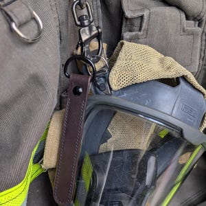 May include: A brown leather strap with a black metal clasp and key ring is attached to a firefighter's mask. The mask is black and gray with a clear visor. The strap is attached to a silver metal clip.