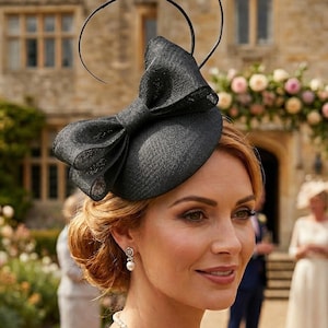 May include: A black bow fascinator with a circular design, worn by a person with auburn hair. The fascinator is made of a textured material and is worn at a wedding. The person is wearing a pearl necklace and earrings.
