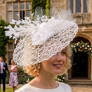May include: A white, open-weave hat with a floral and feather accent. The hat is worn by a person in a white dress, with a blurred background of a garden and a stone building. The hat is suitable for a special event.