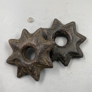 Puede incluir: Dos objetos en forma de estrella de color marrón oscuro con un agujero central, posiblemente decorativos o funcionales. Los objetos tienen una superficie rugosa y texturizada y parecen estar hechos de piedra o un material similar. También hay un pequeño disco plateado.