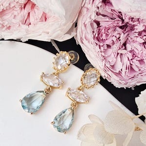 May include: Gold-toned drop earrings with clear and blue-green gemstones. The earrings feature a teardrop-shaped blue-green stone at the bottom, with a clear oval and round stone above. The earrings are set against a backdrop of pink flowers.