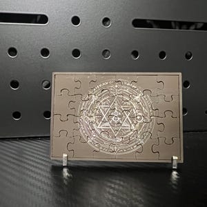 Puede incluir: Un rompecabezas marrón con un diseño geométrico detallado en color plata. El rompecabezas se muestra sobre un soporte, mostrando la imagen completa. El fondo presenta un panel oscuro perforado.