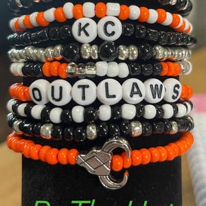 May include: Stack of beaded bracelets in orange, black, white, and silver. Some bracelets spell out "KC" and "OUTLAWS" with white letter beads. The bottom bracelet has a silver charm. The text "Be The Hat" is at the bottom.