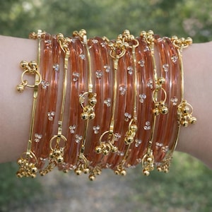 Peut inclure: Ensemble de bracelets de couleur orange avec des détails dorés et de petites clochettes dorées pendantes. Chaque bracelet est orné de minuscules strass étincelants. Les bijoux sont présentés sur un fond vert flou.
