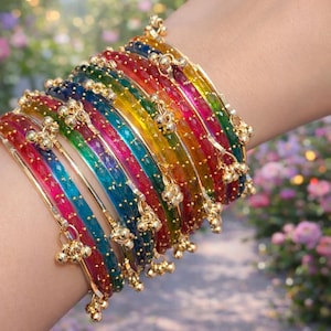 Puede incluir: Una colección de pulseras coloridas en tonos arcoíris, incluyendo rojo, azul, verde y amarillo. Cada pulsera está adornada con pequeños abalorios dorados. Las pulseras están apiladas en una muñeca, creando una exhibición vibrante y llamativa.