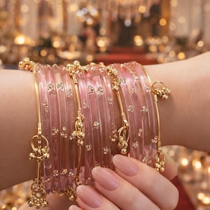 Può includere: Una mano indossa diversi braccialetti rosa con dettagli dorati e ciondoli pendenti. I braccialetti trasparenti presentano piccoli elementi decorativi. Lo sfondo sfocato suggerisce un'ambientazione festosa.