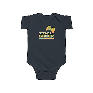 Puede incluir: Body de bebé azul marino con un diseño gráfico de un mando de videojuegos y el texto "TINY GAMER". El diseño utiliza colores amarillo, naranja, verde y morado, ideal para los pequeños jugadores.