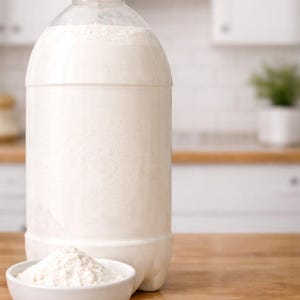 May include: A large, clear plastic bottle with a yellow cap, filled with a white substance, likely flour or a similar ingredient. A small white bowl holds a pile of the same white substance, placed on a wooden surface.
