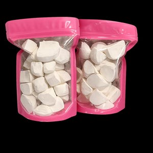 May include: Two stand-up pouches with bright pink plastic edges and clear front windows. Each pouch is filled with numerous irregular, matte white chalk-like chunks. The pouches feature a resealable top closure and are positioned side by side.