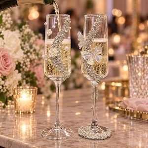 May include: Two clear glass champagne flutes filled with sparkling liquid. The left flute features white pearl accents and small white roses. The right flute is adorned with a silver butterfly and crystal embellishments on the base and stem.