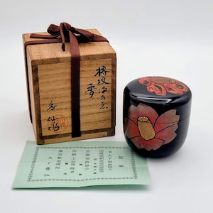 Puede incluir: Una caja de madera con texto japonés y una cinta marrón, junto a un recipiente de laca negra con un diseño floral rojo y dorado. También hay un papel verde con texto japonés.