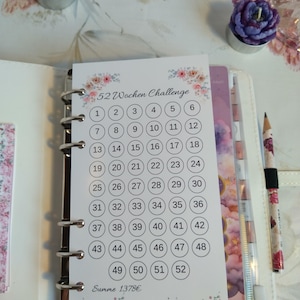 Könnte beinhalten: Ein weißer Planer mit einem 52-Wochen-Challenge-Einsatz. Der Einsatz hat ein Blumendesign und nummerierte Kreise. Zwei lila blütenförmige Kerzen und ein floraler Bleistift sind ebenfalls sichtbar. Der Text "52 Wochen Challenge" steht oben.
