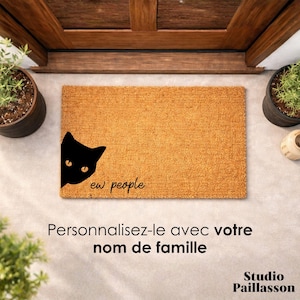 Peut inclure: Un paillasson rectangulaire en fibres naturelles avec un motif de chat noir et le texte "ew people". Le paillasson est placé devant une porte en bois. Des plantes en pot sont visibles.