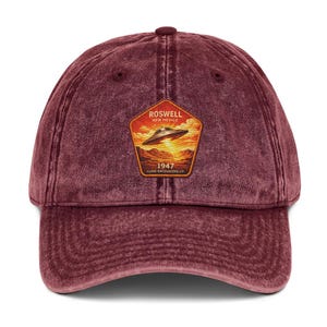 Könnte beinhalten: Weinrote Baseballkappe mit Used-Look. Die Kappe hat einen Aufnäher mit einem UFO über einer Wüstenlandschaft, dem Text "Roswell New Mexico 1947 Close Encounters Co."