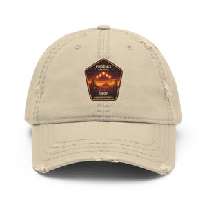 May include: A distressed beige baseball cap featuring a patch with the text "PHOENIX ARIZONA 1997" and a desert landscape with three bright lights. The cap has a pre-worn look with distressed edges.