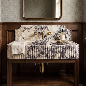 May include: A marble sink with a white and purple veined pattern, set on a dark wood vanity. The sink has gold fixtures and a matching mirror above. A white towel with "MMG" is draped on the side.